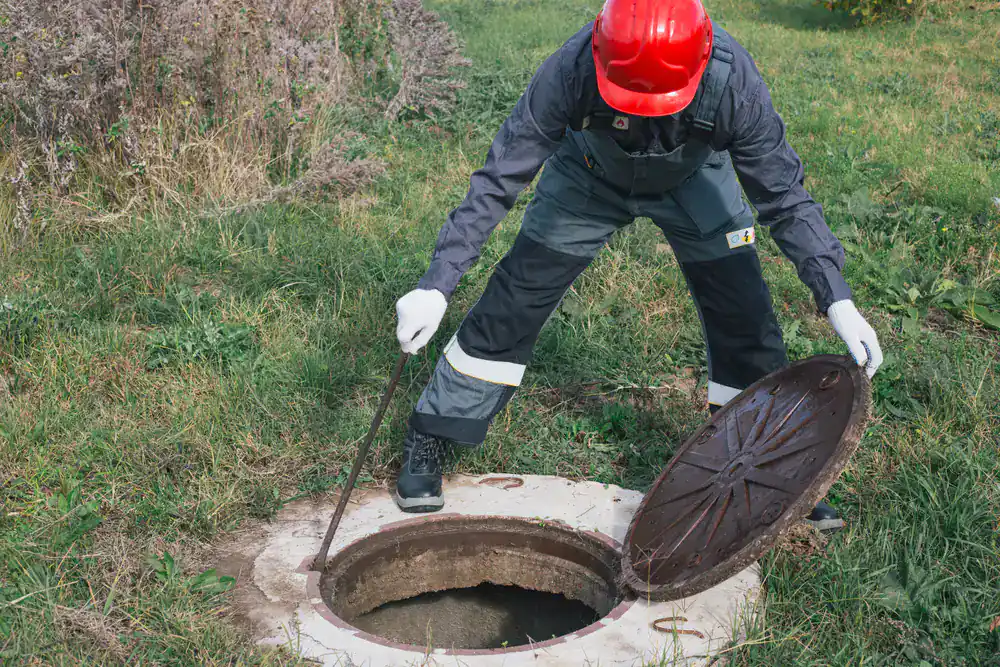 Worker in red helmet lifts a round manhole cover during cesspool service in grassy Suffolk County area.