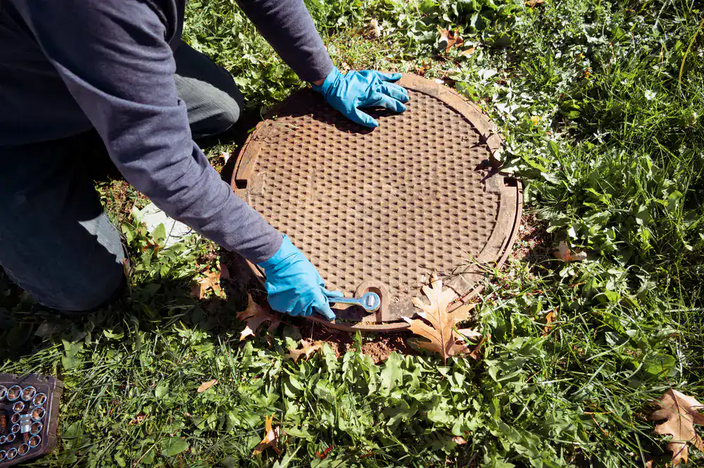 A person in blue gloves kneels on grass in Suffolk County, NY, using a wrench to open a cesspool cover.