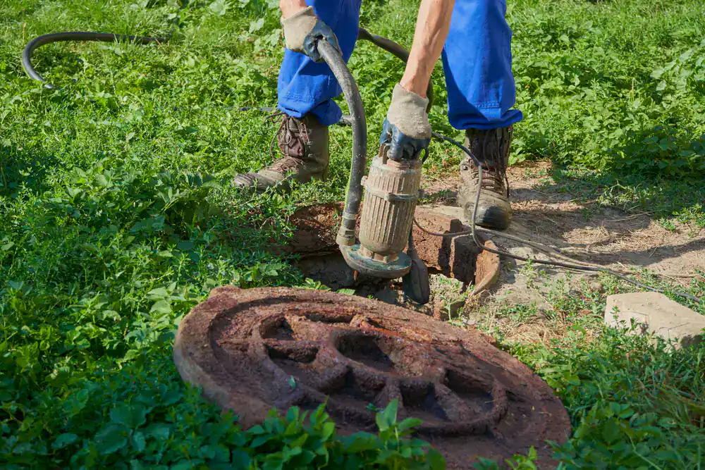 A gloved worker from Septic Services Suffolk County uses a pump beside an open manhole on grass.