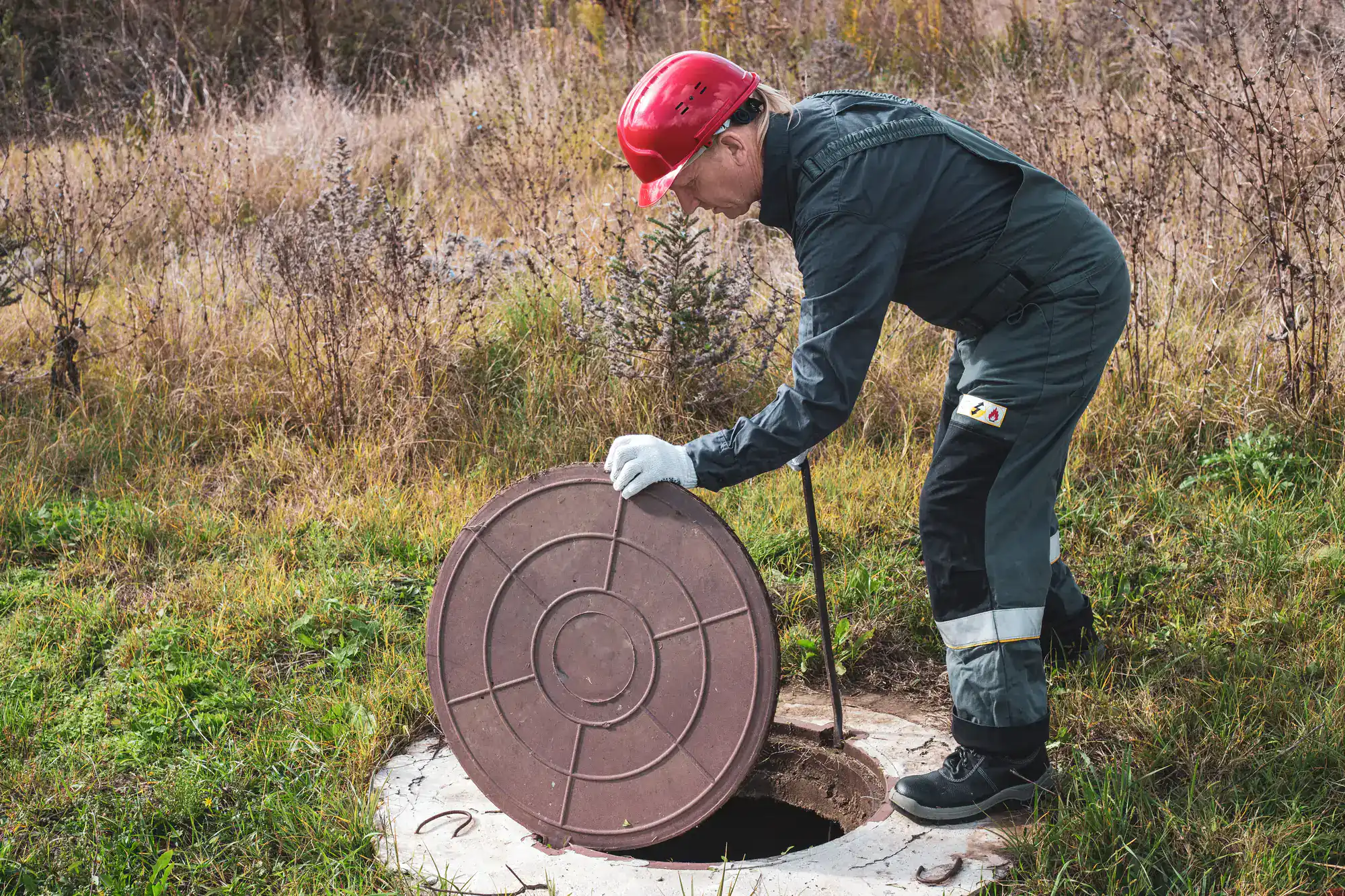A worker in coveralls and a red helmet lifts a round manhole cover outside in a grassy area.